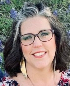 Jennifer Ramirez smiling lokoing at the camera with red glasses, and gold earings on in front of a bluebonnet field.