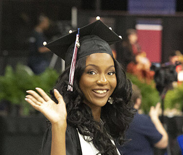 An A&M-Central Texas graduate in academic regalia waving to the camera with a smile on her face.