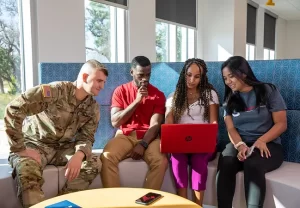 Four students sitting on a bench, looking at a laptop together.