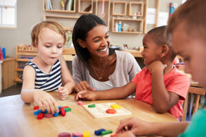 Three kids playing with blocks at a table, with a teacher encouraging them.