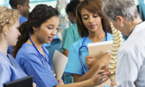 A group of nurses stand in a circle, as one of them points something out on a human spine model.