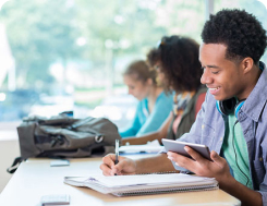 Three college students sit at a desk and work on paperwork. The male student closest to the camera is smiling.