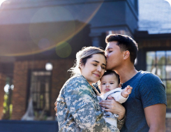 A man and a military-affiliated woman stand in front of a building, with the woman embracing her baby.