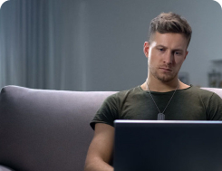 A man looks at a laptop while sitting on a couch. The man has a stern look on his face.