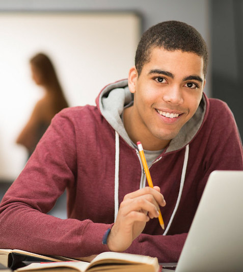 A high school student holding a pencil at a desk with a laptop and notebooks