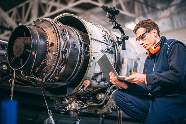 Aircraft engineer in a hangar holding laptop computer while repairing and maintaining a jet engine.