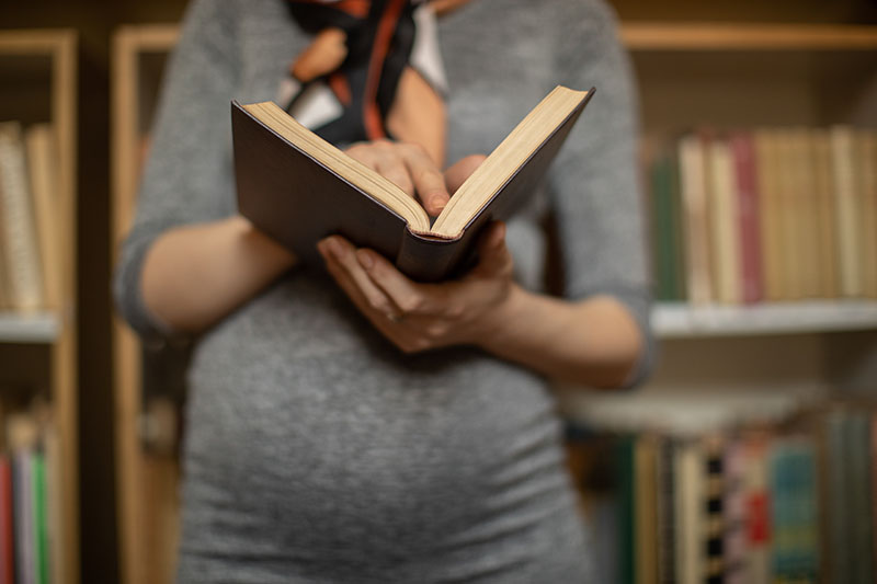 Young pregnant woman holding old book while standing in library in front of bookshelves. She wears tight dress and looks beautiful