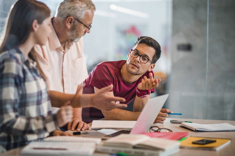 A man leans on a desk while talking to two other people