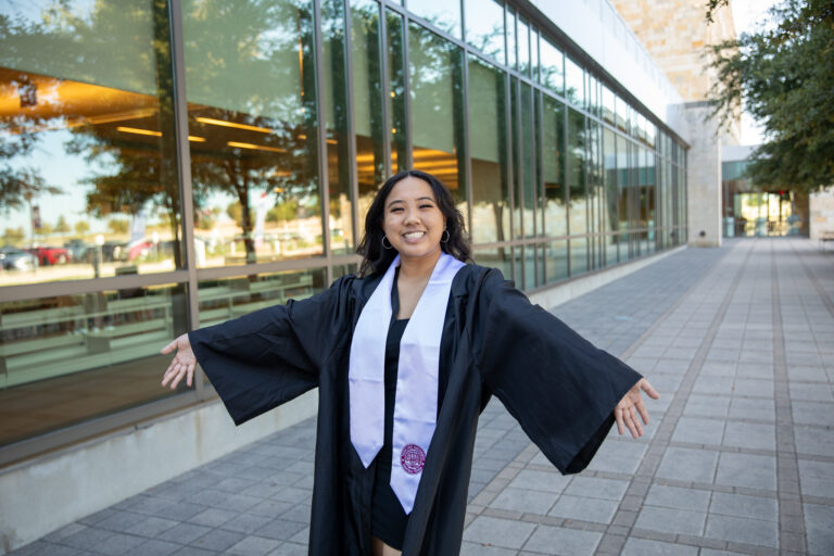 TAMUCT graduate in black gown and white stole smiles in front of the unviersity library