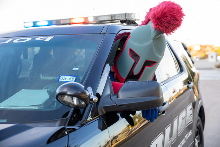Pathfinder mascot in the driver's seat of a TAMUCT police car and poking his head outside of the window.