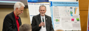 Dr. Rhodes sitting in front of a group, one is holding a microphone and talking.
