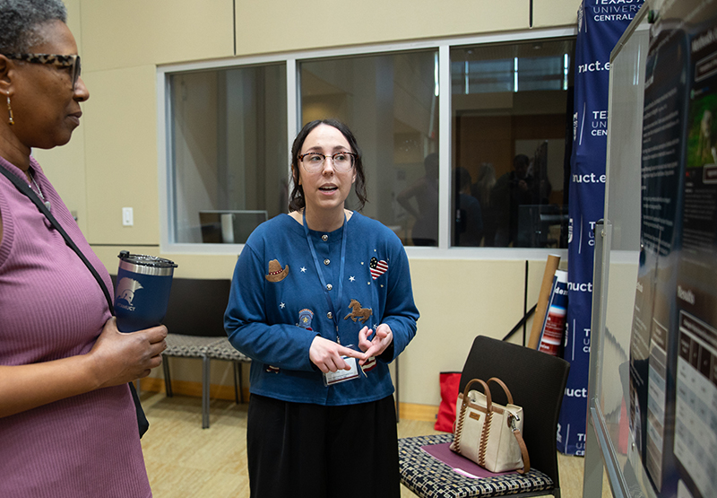 Woman in center of photo explains her research poster to another woman.