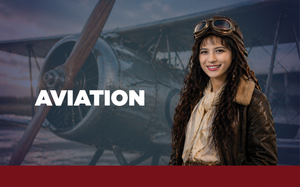 Woman in vintage aviation flight suit in front of a single prop plane with the word "Aviation" displayed.