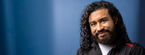 Long-haired man smiling for the camera with a blue background behind him.