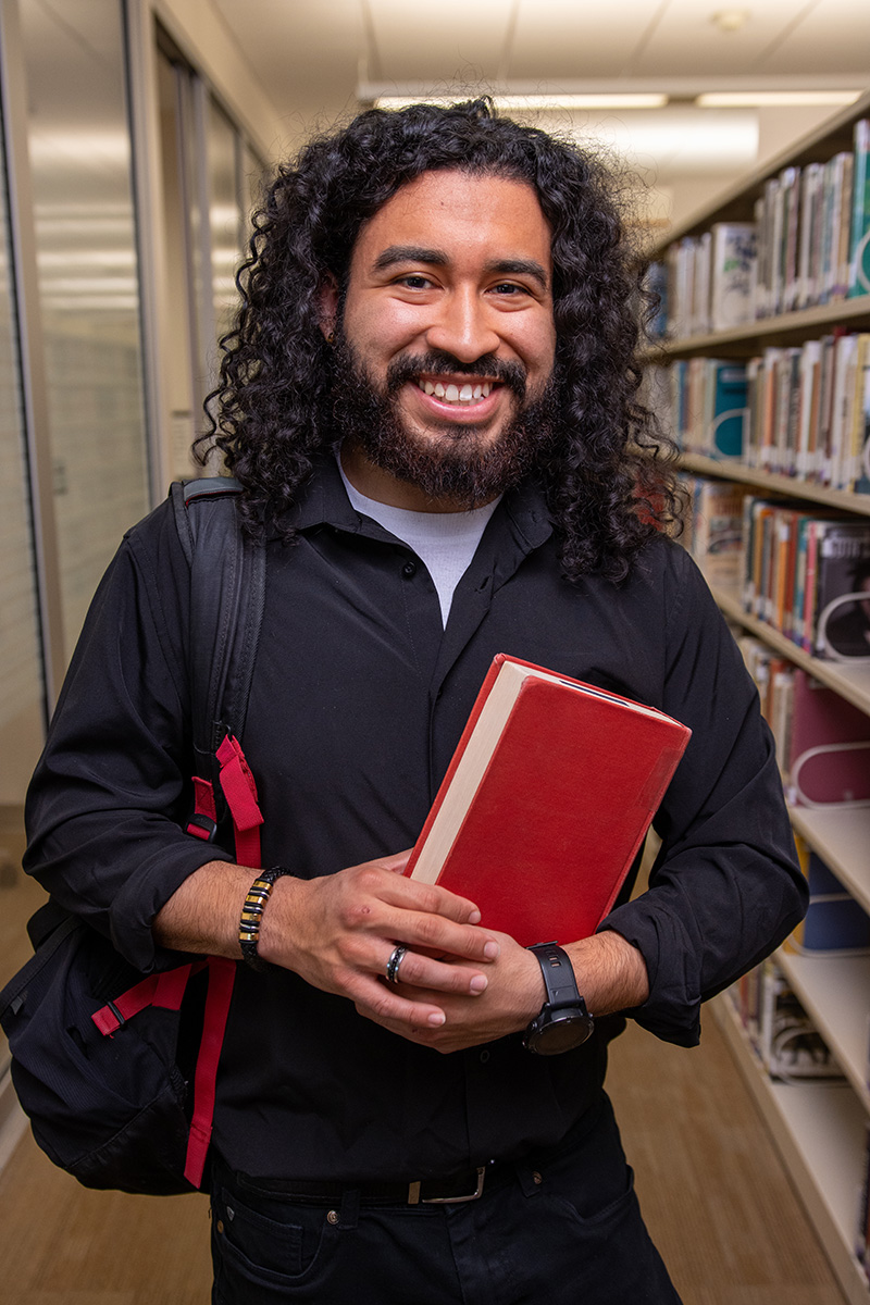 Long-haired man standing in the library holding books and a backpack.