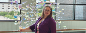 Woman in dress, dark sweater and multi-colored framed glasses stands in front of a cascading butterfly display.