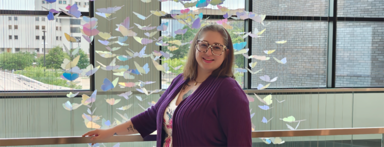 Woman in dress, dark sweater and multi-colored framed glasses stands in front of a cascading butterfly display.
