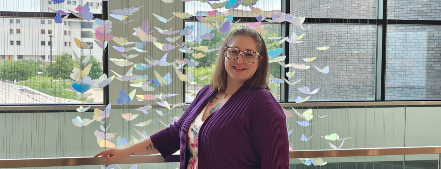 Woman in dress, dark sweater and multi-colored framed glasses stands in front of a cascading butterfly display.