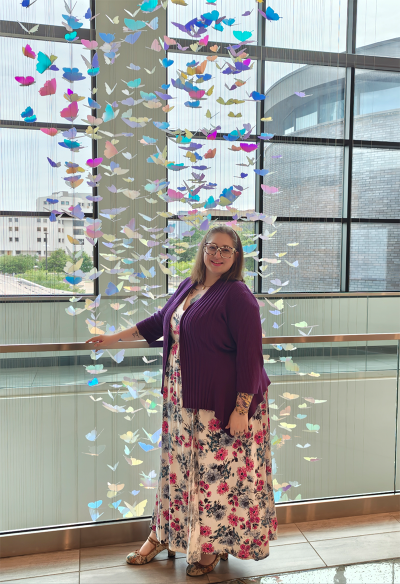 Woman in dress, dark sweater and multi-colored framed glasses stands in front of a cascading butterfly display.