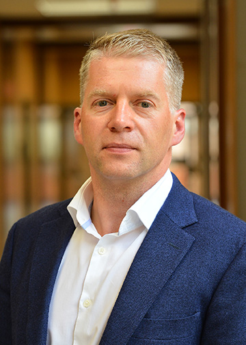 Caucasian man with short blonde hair wearing a white button down shirt and blue sport coat.