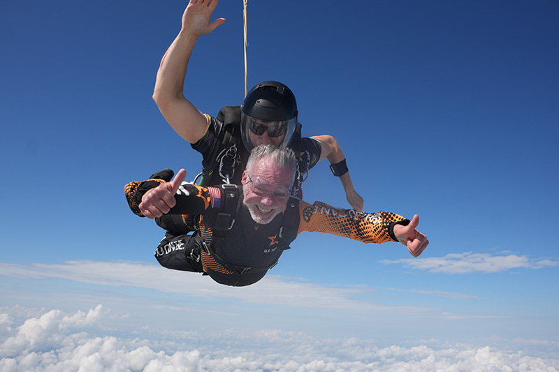 75 year old man gives a thumbs up as he free-falls during a tandem skydiving jump.