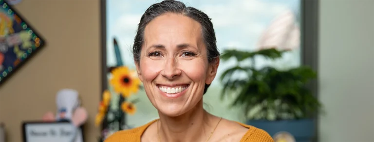 Portrait of a woman in a burnt orange blouse sitting in her office, smiling at the camera.