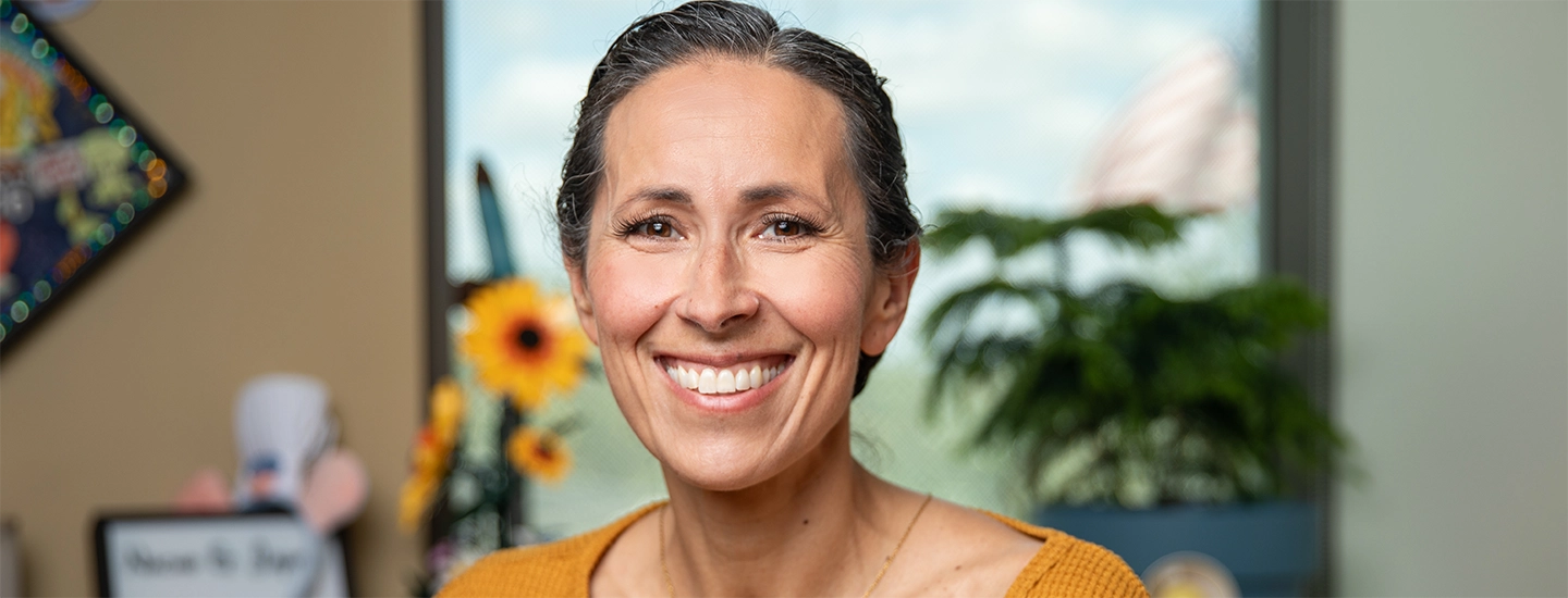 Portrait of a woman in a burnt orange blouse sitting in her office, smiling at the camera.