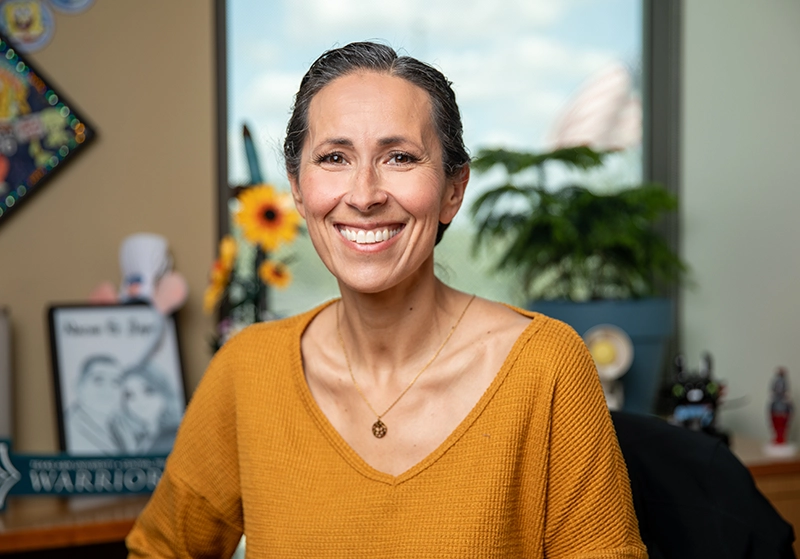 Portrait of a woman in a burnt orange blouse sitting in her office, smiling at the camera.