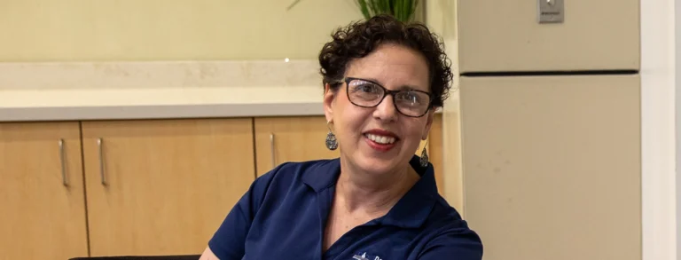 Dr. Laura Erlandson poses for a photo while sitting at a table wearing glasses and blue polo shirt.