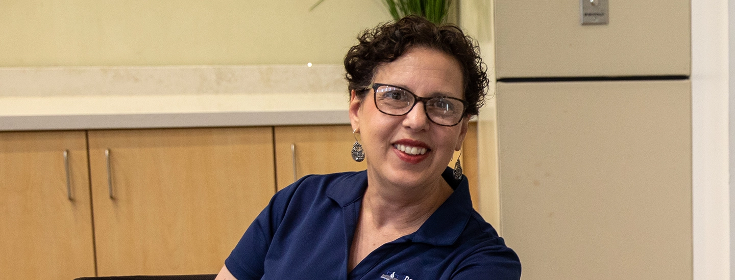 Dr. Laura Erlandson poses for a photo while sitting at a table wearing glasses and blue polo shirt.