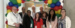 Four men and five women stand as a group under a multi-colored balloon arch.