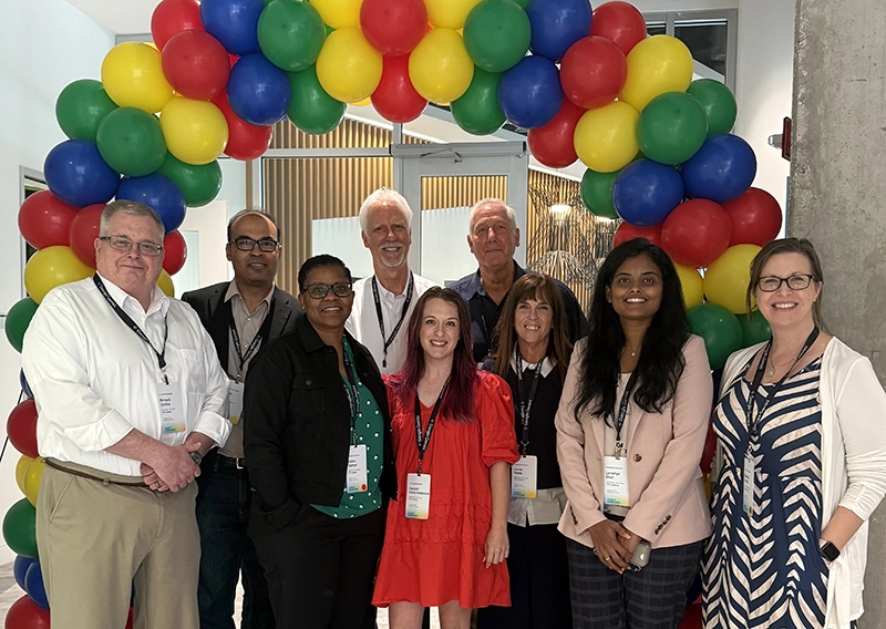 Four men and five women stand as a group under a multi-colored balloon arch.