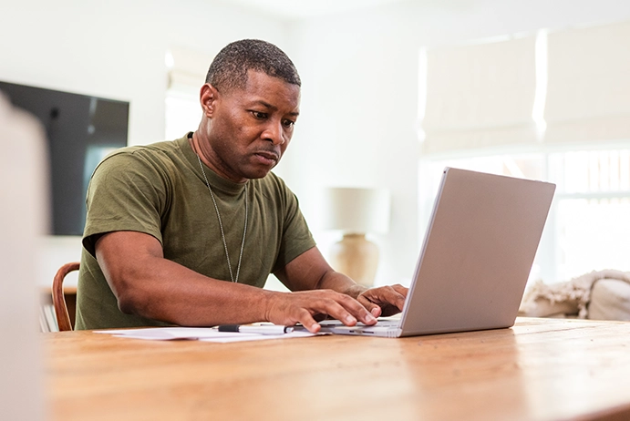 Black man in an Army green t-shirt sitting at a table working on a laptop.