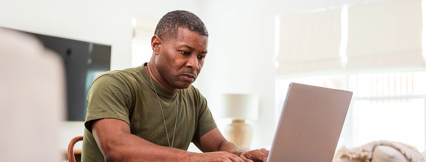 Black man in an Army green t-shirt sitting at a table working on a laptop.