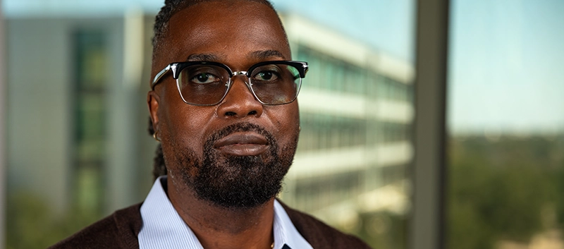 Headshot of a black man wearing glass with the TAMUCT campus behind him.