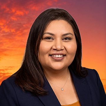 Woman with long, dark, hair, smiling for the camera, sitting in front of a sunset background.