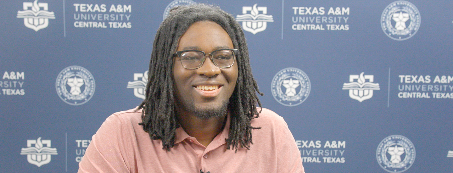 Black man with dreadlocks, wearing glasses, sitting in front of a step-and-repeat banner for the university.