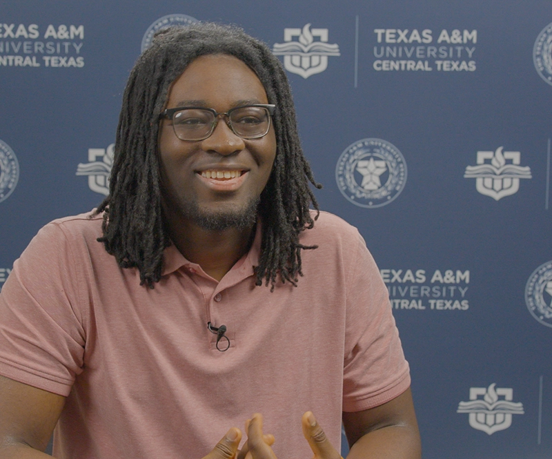 Black man with dreadlocks, wearing glasses, sitting in front of a step-and-repeat banner for the university.