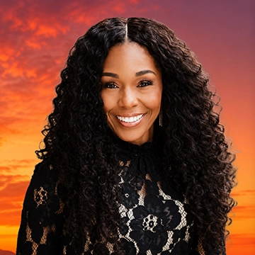 Black woman with long hair, smiling at the camera, sitting in front of a sunset background.