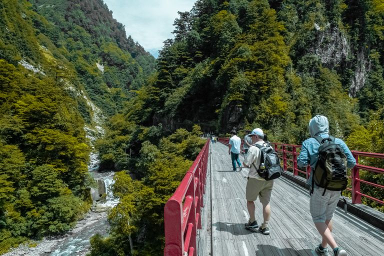 Hikers crossing a red bridge in a lush mountain landscape under daylight.