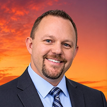 Man with short, cropped hair, and a goatee and mustache, smiling at the camera, sitting in front of a sunset background.
