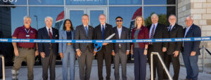 Ten people stand in a line behind a "grand opening" ribbon. The man in the center is holding a large pair of scissors.
