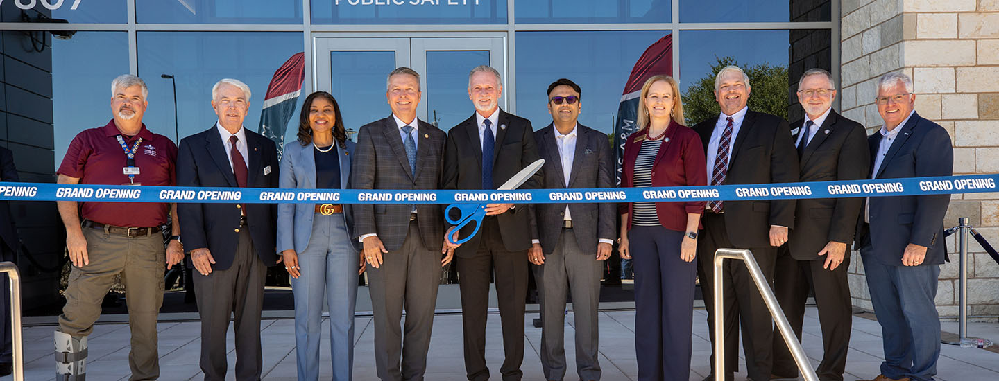 Ten people stand in a line behind a "grand opening" ribbon. The man in the center is holding a large pair of scissors.