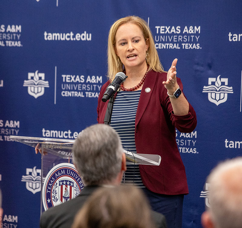 Blond haired woman in a maroon blazer stands at a podium and address the crowd.