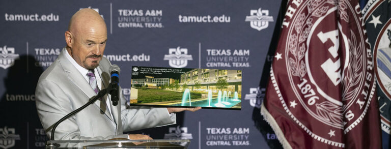 Bald man in a grey suit holds an image of a university building while speaking into a microphone.