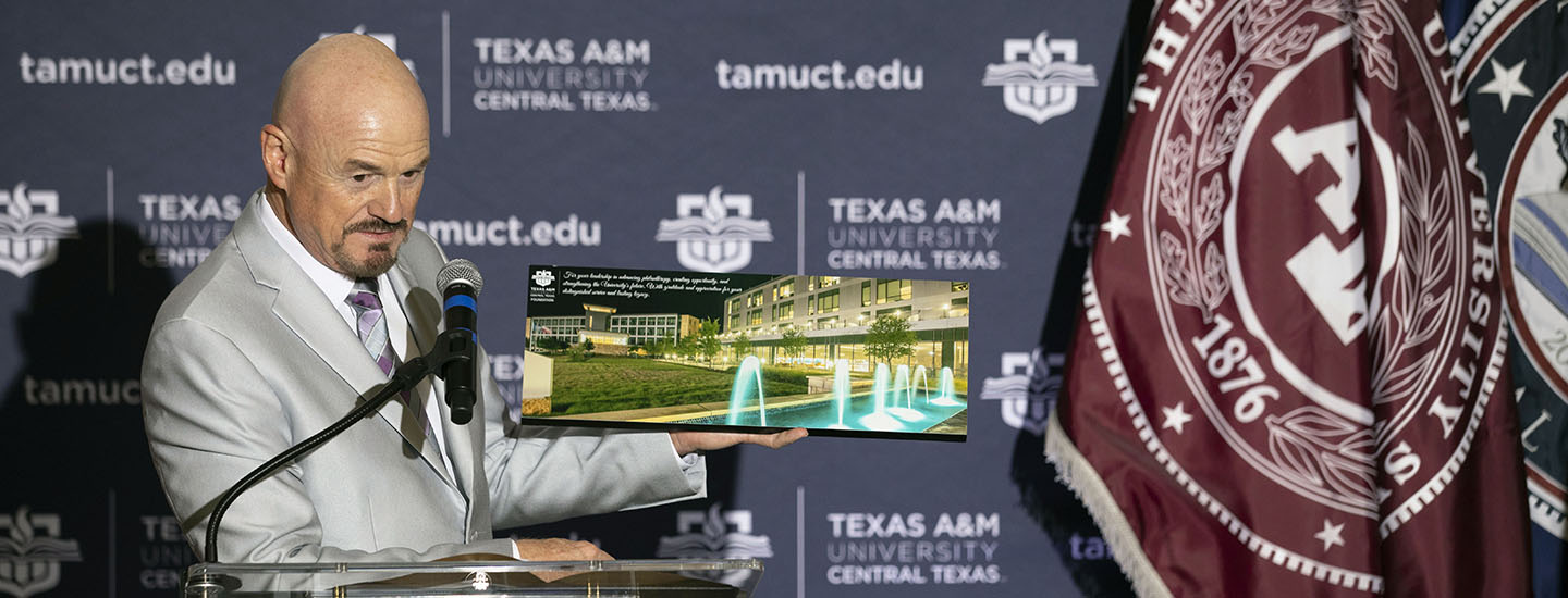 Bald man in a grey suit holds an image of a university building while speaking into a microphone.