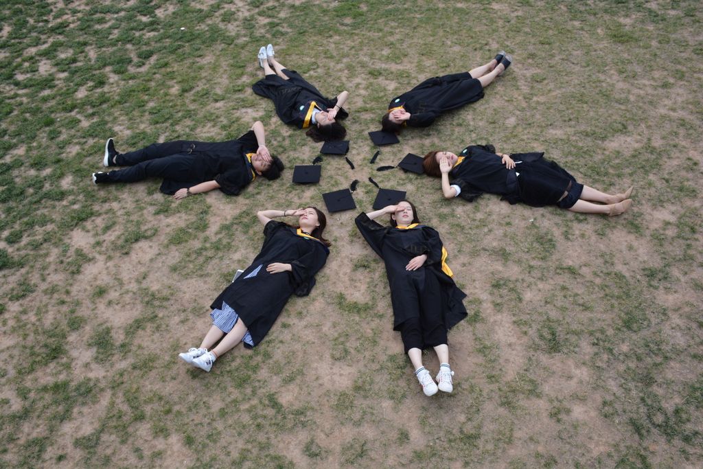Overhead view of six graduates lying in circle on grass, celebrating.