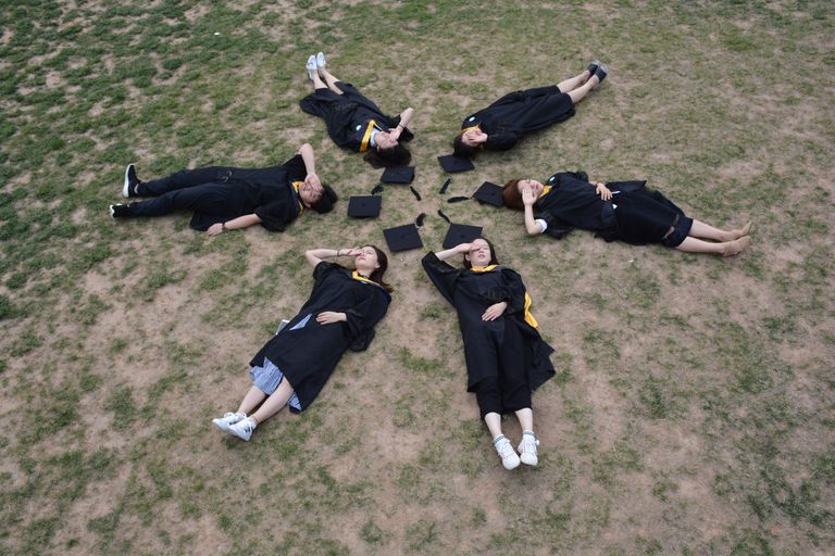 Overhead view of six graduates lying in circle on grass, celebrating.