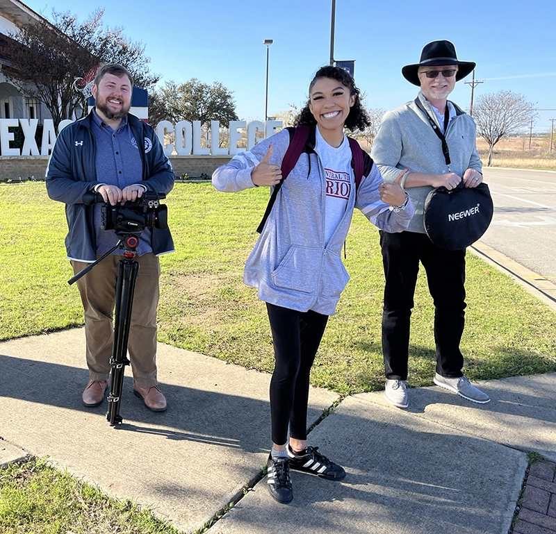 Young woman stands between two men giving two thumbs up in front of the Central Texas College sign. The men are carrying camera equipment.